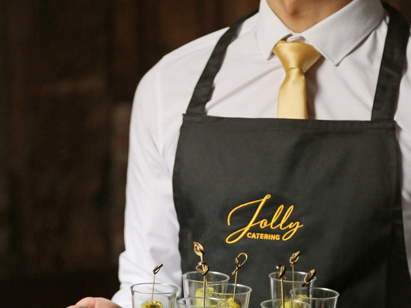 Catering staff member in an embroidered Jolly Catering apron, white hospitality shirt and gold tie serving canapes