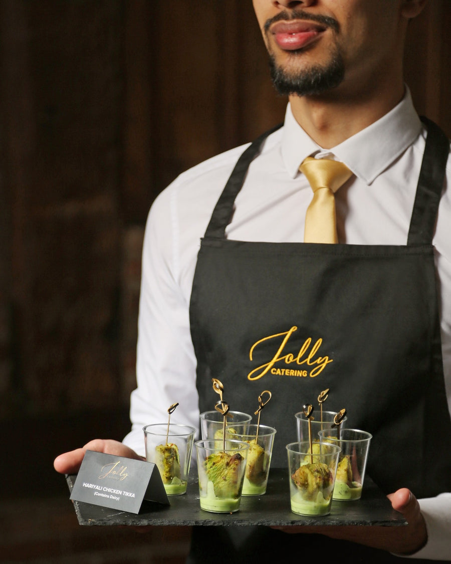Catering staff member in an embroidered Jolly Catering apron, white hospitality shirt and gold tie serving canapes