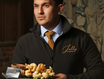 Jolly Catering team member in a black zipped jacket with a gold embroidered logo, carrying a platter of canapes