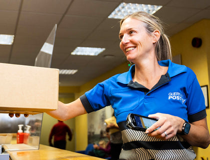 Guernsey Post counter colleague in a branded blue polo shirt handing a parcel across the counter