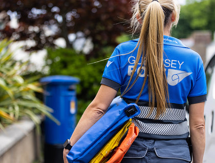 Guernsey Post postie photographed from behind, showing the reflective printed back logo and hi-vis banding on the delivery polo